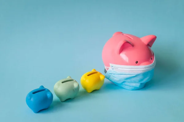 Tıbbi maskeli ve domuz yavrusu kumbaralı domuz kumbarası. Koruyucu maske takan pembe domuzcuk kumbarası ve izole edilmiş mavi arka planda küçük kumbaraları takip ediyor. Koronavirüs salgınında para tasarrufu.