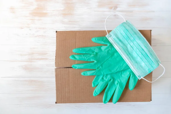 Green rubber gloves and medical face mask on closed donation carton box. Protective facemask and gloves on cardboard box for donation. Top view with copy space. Sponsorship tradition concept.