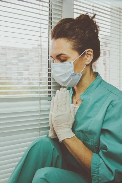 Tired Doctor woman in medical mask, gloves and doctors suit closed her eyes, clasped her hands in a gesture of Namaste and praying.