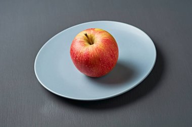 Vitaminlerle dolu tek bir kırmızı taze elma yuvarlak seramik tabakta ve mutfaktaki eski koyu çizik beton masada yatıyor. Hasat ya da diyet konsepti. Yakın plan.