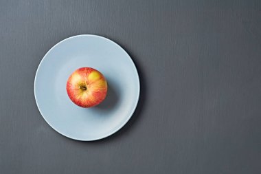 Vitaminlerle dolu tek bir kırmızı taze elma yuvarlak seramik tabakta ve mutfaktaki eski koyu çizik beton masada yatıyor. Hasat ya da diyet konsepti. Mesaj için yer var. Üst görünüm