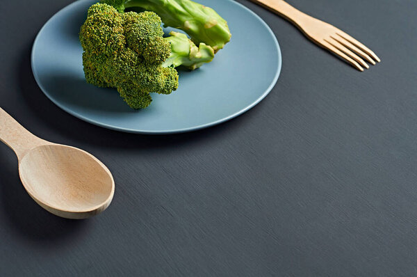 Two bunches of broccoli in gray ceramic plate near wooden spoon and fork lies on scratched dark concrete desk on kitchen. Space for text