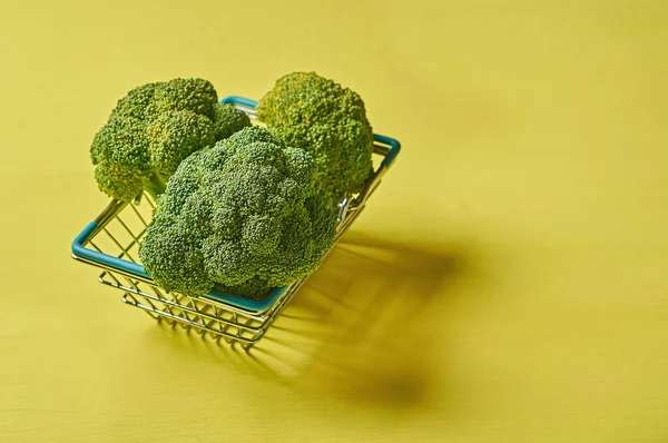 Metal market basket full of bunches of broccoli lies on scratched yellow concrete desk on kitchen. Space for text