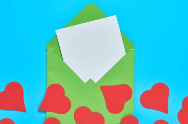 Açık yeşil posta zarfının yanında kırmızı kalpler ve mavi masada boş beyaz kağıt. Aşk mesajı. Sevgililer Günü konsepti. Mesaj için yer var. Üst görünüm