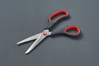 Plastik saplı tek keskin makas siyah ve kırmızı renkler koyu beton masanın üzerinde yatar. Yakın plan.