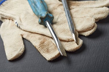 Tamir ya da diğer zor işler için bir çift kirli tekstil koruyucu eldiven plastik saplı eski dağınık tornavidaların yanında çalışma odasındaki koyu çizik beton masada yatıyor. Yakın plan.