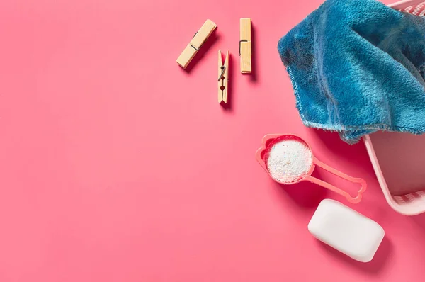 Plastic basket for washing with soft towel near antibacterial soap, clothes pin and measuring spoon full of powder lies on pink countertop in laundry. Space for text. Top view