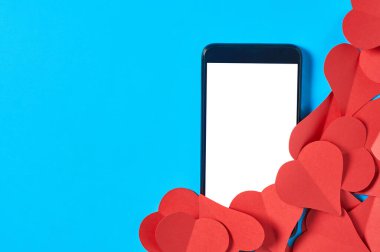 Siyah akıllı telefon, mesaj, resim, fotoğraf ve diğer grafikler için izole edilmiş beyaz ekranlı, mavi masadaki kırmızı kağıt kalpler yığınının yanında. Sırlar ya da yasak aşk kavramı. Mesaj için yer var. Üst görünüm