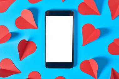 Mesajlar, resimler, fotoğraflar ve dağınık kırmızı kağıt kalplerin yakınındaki diğer grafikler için izole edilmiş beyaz ekranlı siyah akıllı telefon mavi masada yatıyor. Sevgililer Günü ve aşk konsepti. Mesaj için yer var. Üst görünüm