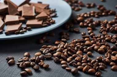 Mavi yuvarlak seramik tabakta bir yığın kırık acı ya da süt çikolatası ve dağılmış kahve çekirdekleri koyu çizik beton masanın üzerinde yatıyor. Yakın plan.