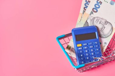 Ukrayna Hryvnia banknotu ve mavi hesap makinesi. Pembe arka planda metal market sepeti. Satın al ya da sat. Döviz kurları kavramı