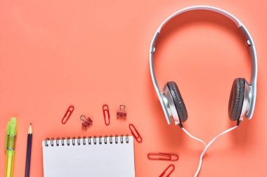 Kablolu, boş kağıt defter ve turuncu arka planda diğer kırtasiye malzemeleriyle modern beyaz kulaklıklar. İş ya da okul konsepti. Uzayı kopyala Üst görünüm