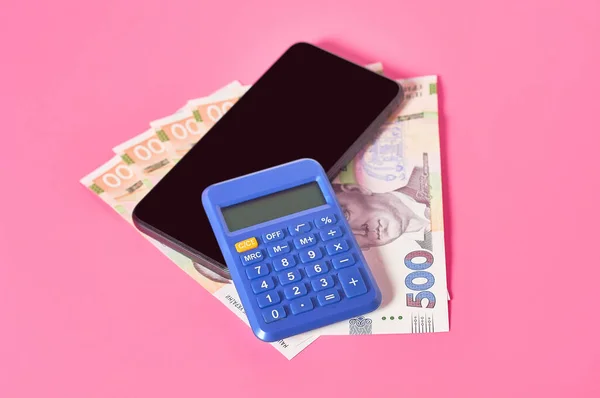 Smartphone, calculator and ukrainian money on pink background. Concept of mobile banking. Online shopping from home. Distancing control financial balance