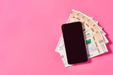 Pembe arka planda akıllı telefon ve Ukrayna parası. Mobil bankacılık kavramı. Evden online alışveriş. Uzaklık denetimi mali denge