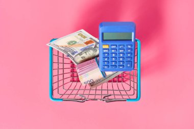 Farklı banknotlar ve pembe arka planda metal market sepetinde mavi hesap makinesi. Satın al ya da sat. Döviz kurları kavramı