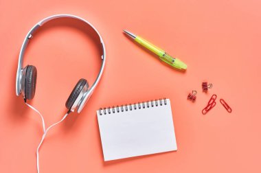 Kablolu, boş kağıt defter ve turuncu arka planda diğer kırtasiye malzemeleriyle modern beyaz kulaklıklar. İş ya da okul konsepti. Uzayı kopyala Üst görünüm