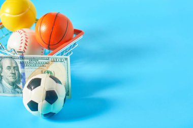 Farklı toplar ve 100 dolarlık banknot mavi arka planda metal market sepeti. Spor aksesuarları alıyorum. Sporda yolsuzluk kavramı