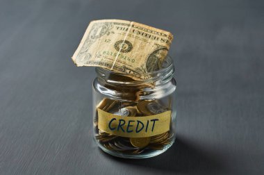 Bozuk paralarla dolu cam kavanoz ve yazıt kredisi, bir dolarlık banknot beton masa üzerinde.