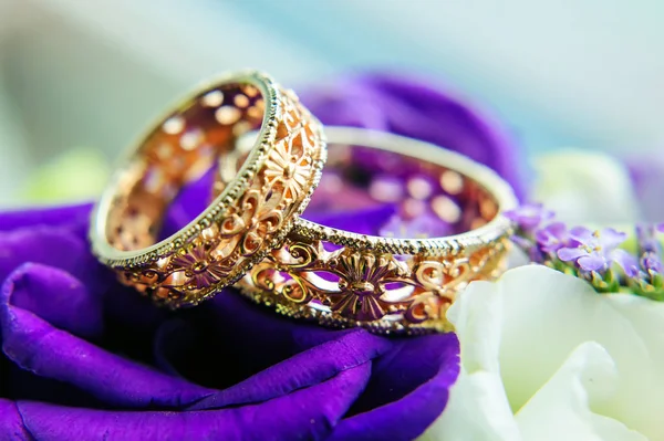 Beautiful gold wedding rings with patterns, close-up. Two gold rings, blurred background with white and blue flowers.
