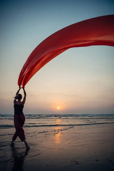 Elegant young girl performs dance moves on the sand holding a long red cloth against the background of evening sky and sun setting over the sea. Silhouette of slim woman against the sunset.