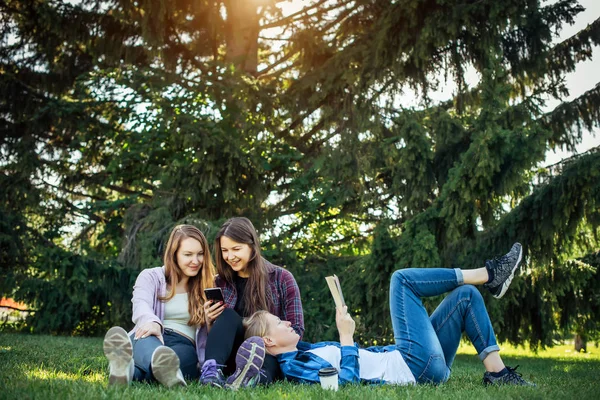 Happy young girls have fun laughing sitting on green grass in the park. Students in between lectures. Blonde, brunette and red-hair girl communicate, look at smartphone, concept of female friendship.