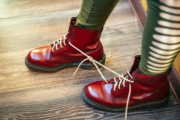 Female legs in stylish bright red boots with thick white laces tied together on wooden background, side view. Woman in funny pants and big size shoes. April Fools\' day, pranks and fun.