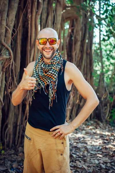 Portrait of stylish young bald guy in sunglasses on blurry background outdoor. Attractive, cheerful man poses for the camera, smiles, and gives thumbs-up.
