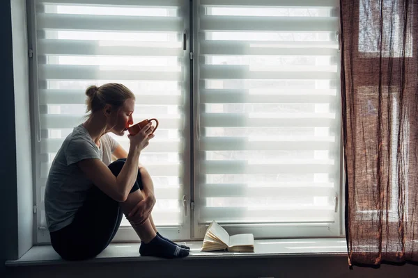 Illness young girl on home quarantine. Woman sits on windowsill, reads a book, drinks tea. Coronavirus pandemic, epidemic in Europe, self-isolation.