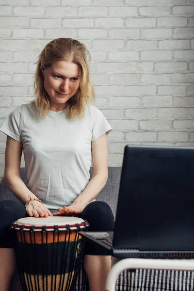 Online music lessons during quarantine due to coronavirus pandemic. Remote teaching to play the drum. Young woman watches video course on playing djembe. Hobbies and leisure activities in lockdown.