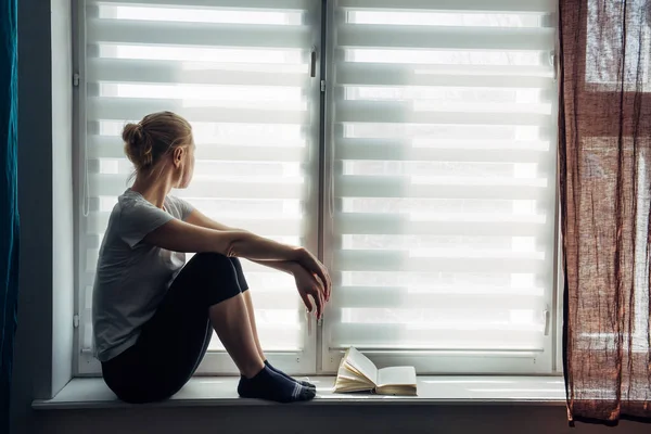 Illness young girl on home quarantine. Woman in protective medical masks sits on windowsill and looks out window. Virus protection, coronavirus pandemic, prevention epidemic, self-isolation.