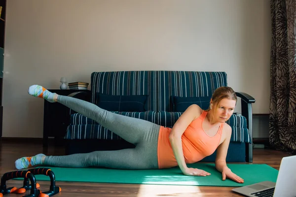 Athletic young woman trains at home during quarantine. Young girl doing fitness exercise for the legs with help of online training programs on laptop. Cute blonde workout on the mat.