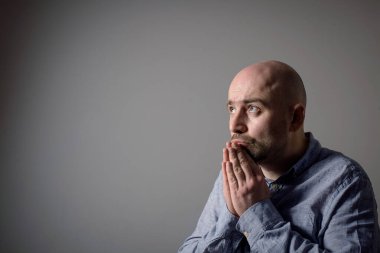 Gri arka planda gri gömlekli, üzgün, kel bir adamın portresi. Yan görünüm. Dua pozisyonu