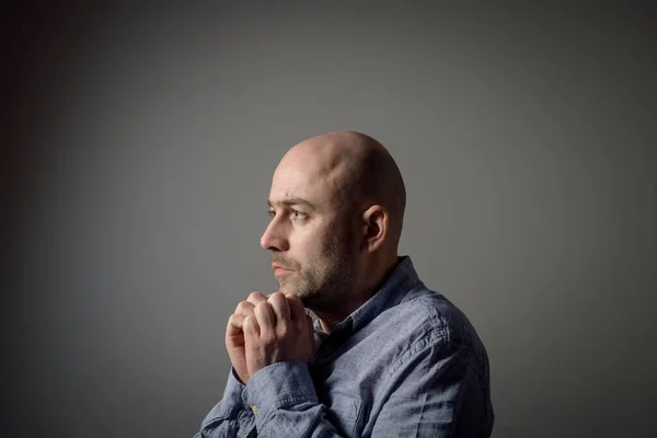 Portrait of caucasian depressed sad bald man in grey shirt on grey background. Side profile view.