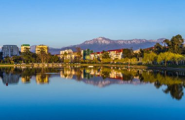 Batum, Gürcistan - 22 Mart 2020. Karlı dağlar, evler ve bir park gölün yüzeyine yansıyor..