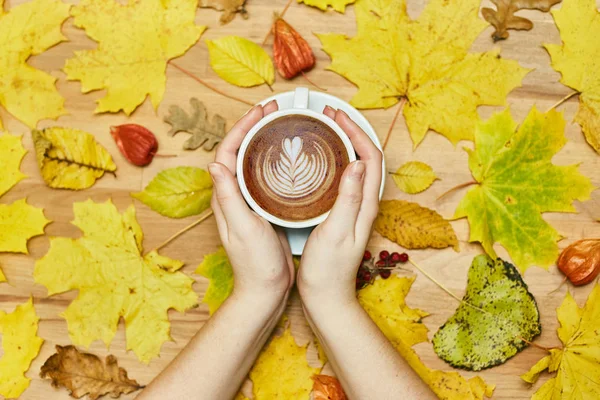 Composición plana de otoño con marco de corona de hojas secas y taza de café con leche en mano de mujer sobre fondo de madera . — Foto de Stock