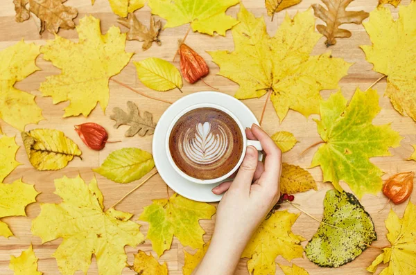 Composición plana de otoño con marco de corona de hojas secas y taza de café con leche en mano de mujer sobre fondo de madera . — Foto de Stock