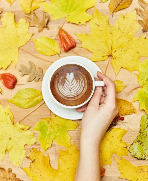 Composición plana de otoño con marco de corona de hojas secas y taza de café con leche en mano de mujer sobre fondo de madera . — Foto de Stock