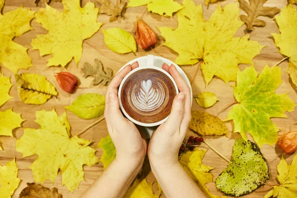 Composición plana de otoño con marco de corona de hojas secas y taza de café con leche en mano de mujer sobre fondo de madera . — Foto de Stock