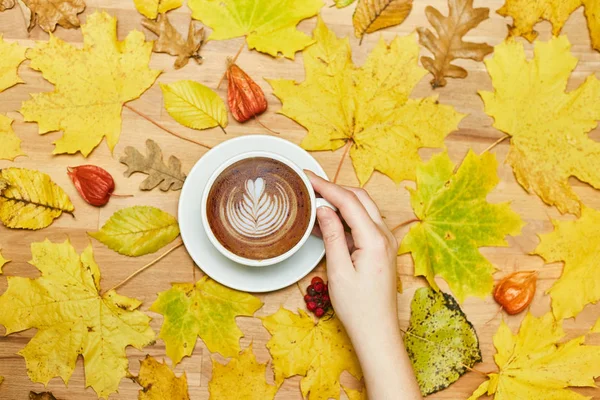 Composición plana de otoño con marco de corona de hojas secas y taza de café con leche en mano de mujer sobre fondo de madera . — Foto de Stock