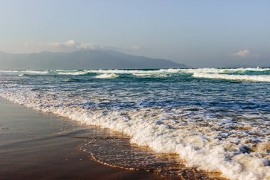 Kumsal kıyısı, denizde sakin yumuşak dalgalar, güneşli hava, beyaz bulutlu mavi gökyüzü. Güzel bir deniz manzarası olan boş, uzun bir sahil. Deniz dalgasından beyaz köpük