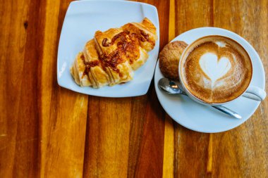Kapuçino kapuçinosu kapuçinosu kapuçinonun içinde bir fincan dolusu kurabiye ve çay tabağında taze kruvasan. Kahve köpüğüne çizilmiş bir kalbin üst görüntüsü. Sabah kahvesi ahşap kahverengi bir masanın üzerinde duruyor. Sabah mükemmel bir kahvaltı, öğle yemeği. kopyalama yeri