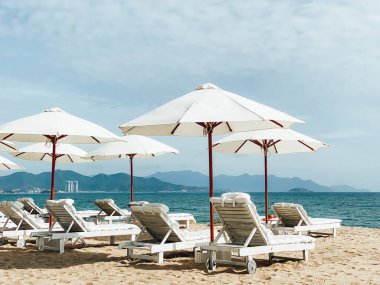 Kumlu bir sahil kıyısındaki beyaz, yumuşak güneş pansiyonları. Tropik tatil köyü, sabah güneşi, mavi gökyüzündeki beyaz bulutlar, deniz manzarası, dağlar ve arka plandaki şehir.