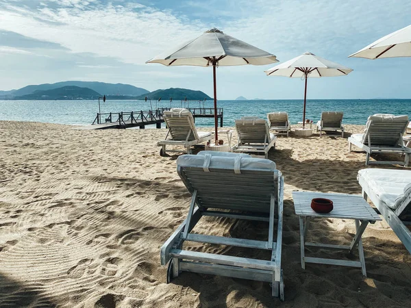 Kumlu bir sahil kıyısındaki beyaz, yumuşak güneş pansiyonları. Tropik tatil köyü, sabah güneşi, mavi gökyüzündeki beyaz bulutlar, deniz manzarası, dağlar ve arka plandaki şehir.