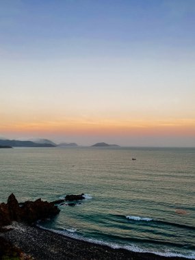 Uçurumun tepesinden panoramik manzaralı gün batımı. Kayalık sahil manzarası, kıyıda dalgalar. Ufuktaki dağlar. Batan güneşten güzel gökyüzü rengi. Güzel dramatik deniz manzarası