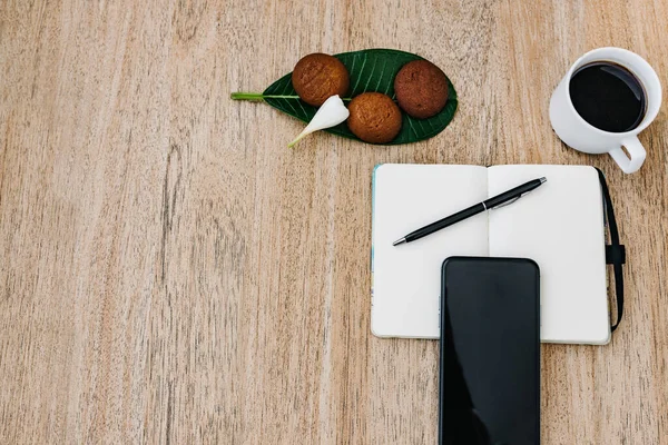 Düz kompozisyon, not defteri, akıllı telefon, bir fincan kahve ve kahvaltı kurabiyesi. O gün için bir plan kaydet. Sabah işyerinde. Açık bir kağıda yazmak için bir kalem. Boşluğu kopyala, metin için boşluk
