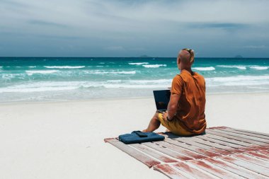 Uzun saçlı, güneşli bir günde deniz kenarında oturan, dizüstü bilgisayarını kullanan, uzaktan kumandalı genç bir adam. Blogcu, serbest çalışan, internet, iş seyahati. Yaz tatili