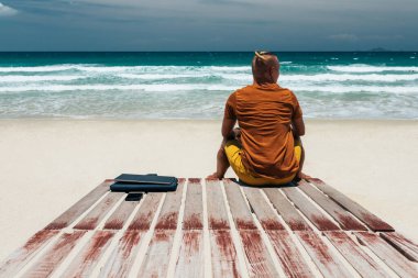 Güneşli bir günde deniz kenarında uzun saçlı genç bir adam sırtı kameraya dönük bir şekilde oturuyor ve yakınlarda bir bilgisayarla iş yapıyor. Blogcu, serbest yazar, iş seyahati. Yaz tatili. Yer kopyala