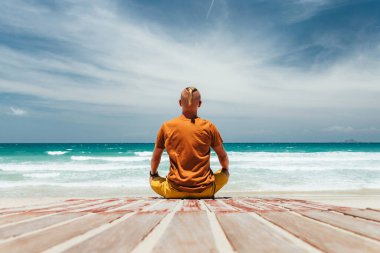 Genç adam tropikal bir plajın kıyısında oturur. Arkasından manzara, meditasyon yeri. Açık bir yaz gününde, kumlu plajlı panoramik deniz manzarası, gökyüzü beyaz bulutlar. Yer kopyala