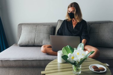 Yüzünde maske olan bir kadın kanepede dizüstü bilgisayarda çalışıyor, karantina süresi, kendini izole etme, evde çalışma. Sabah uyandıktan sonra, sabah kurabiyeli kahve, masada çiçekler.