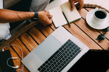 Çalışma alanının dizüstü bilgisayarında çalışan bir adamın elleri, ahşap bir masanın üzerinde, akıllı bir telefonla, bir fincan kahve ve kayıt davaları için bir defter. İş konsepti. Uzaktan kumanda. Boşluğu kopyala, tahta arkaplan